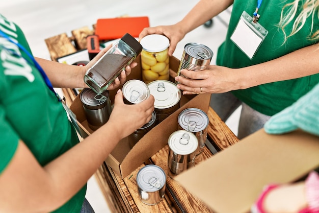 Close-up image of a person's hands donating canned goods to a food bank volunteer, with a shopping cart filled with various food items in the background.