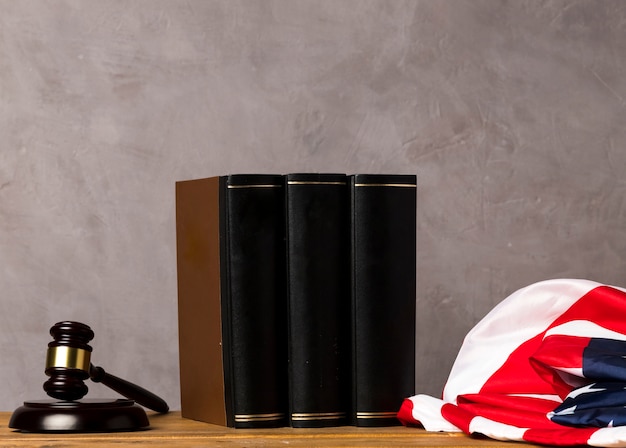 A gavel resting on top of a stack of law books in a courthouse setting. The scene is dimly lit to create a solemn and serious atmosphere. A blurred American flag in the background.