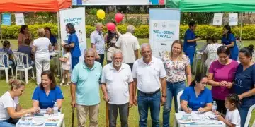 Pessoas de diferentes idades participando de uma feira de saúde comunitária ao ar livre, com profissionais de saúde oferecendo serviços e informações.