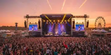Thousands of music fans enjoying a vibrant outdoor festival at sunset, with a band performing on a brightly lit stage.