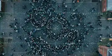 An aerial drone shot of a regional community festival, with hundreds of people forming geometric patterns on cobblestones under overcast skies, highlighting community well-being and social connection.
